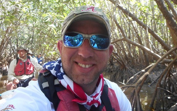 a person in a hat, sunglasses and an american flag bandana smiles from a canoe on an outward bound veterans expedition 